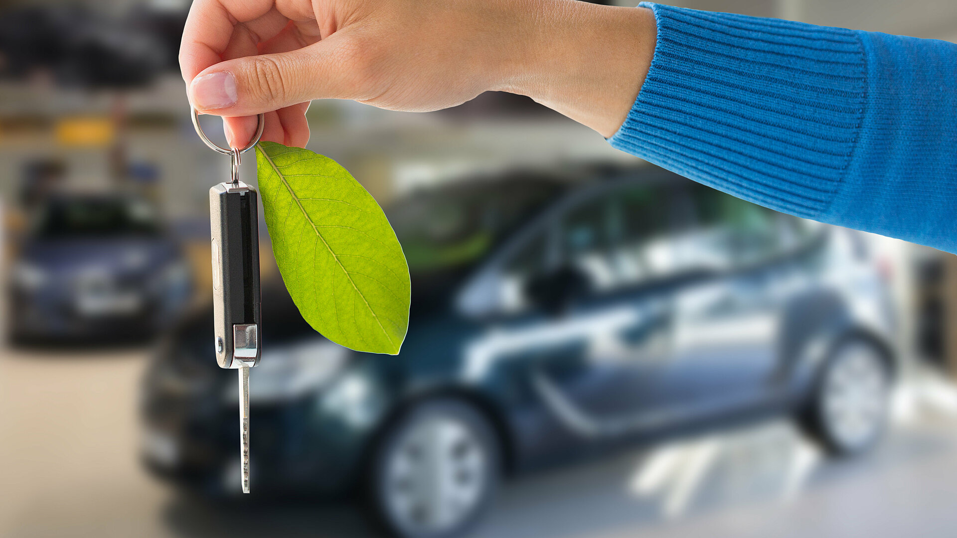 Hand hält Autoschlüssel mit grünem Blatt als Anhänger. Im Hintergund ist unscharf ein Auto zu sehen