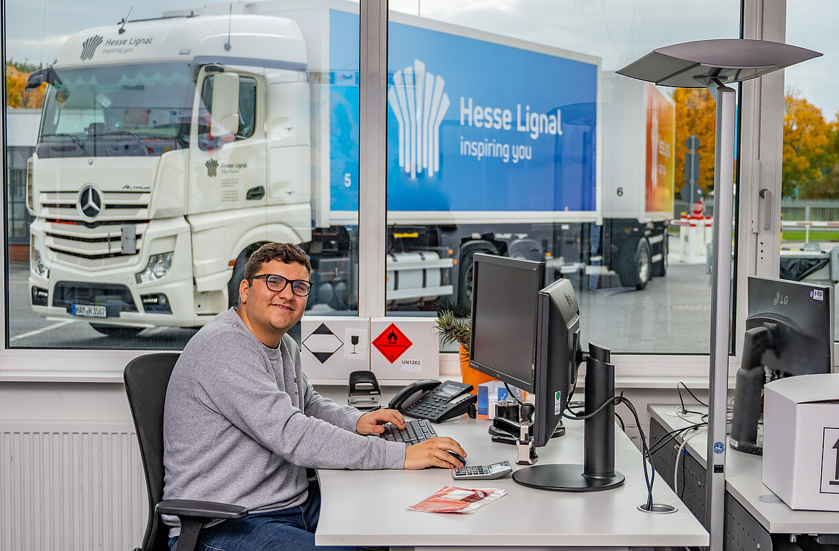 Kauf­frau/-mann für Spe­di­ti­on und Lo­gis­tik­dienst­leis­tun­gen am Schreibtisch und einem Hesse Lignal LKW im Hintergrund